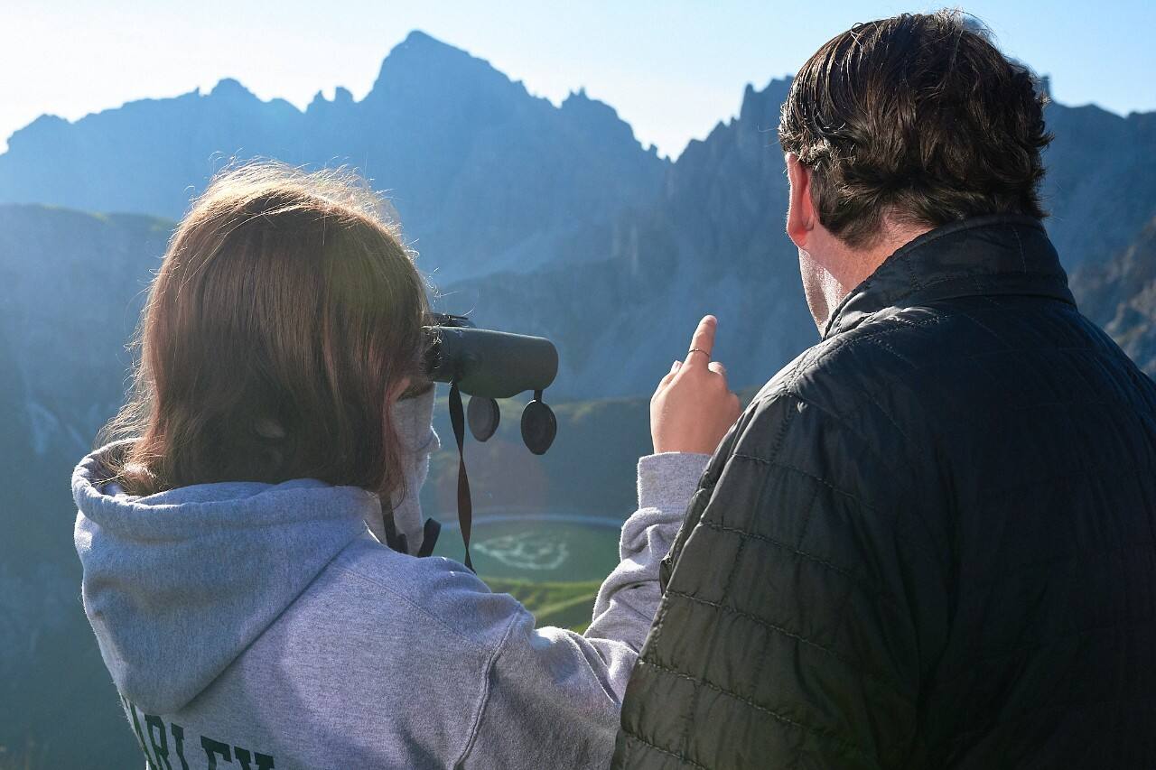 A man and woman look out over a mountain landscape. Seen from the back, the woman is looking through binoculars and pointing, 